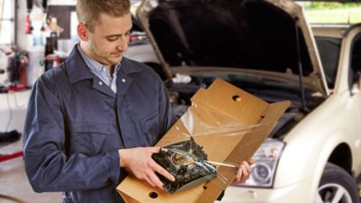 Un mecánico está parado frente a un vehículo con el capó abierto y está abriendo un paquete que contiene una pieza de automóvil.