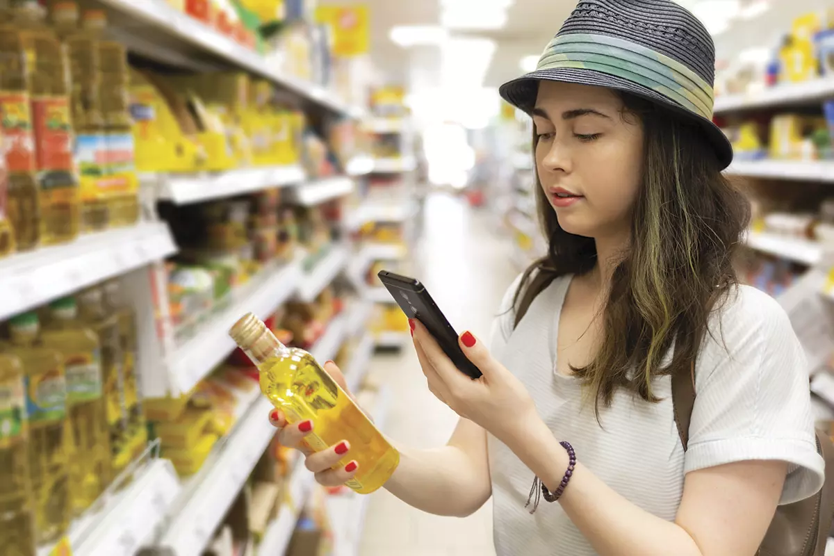 A young girl wearing a hat is scanning the QR code on a bottle of olive oil in a grocery store.